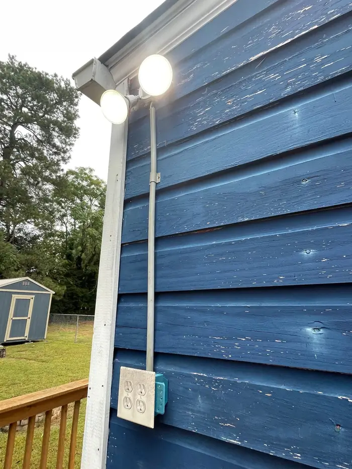 Two exterior floodlights lit on the corner of a house, fed by conduit down to an outdoor receptacle